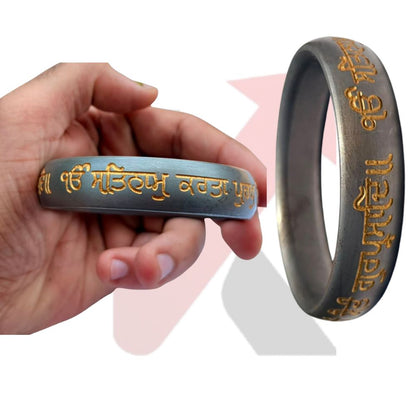 A dark gray metallic bracelet( Punjabi kada)  with gold-colored Punjabi script, shown being held by a hand on the left and as a larger standalone view on the right against a white background with soft pink and gray graphic shapes.