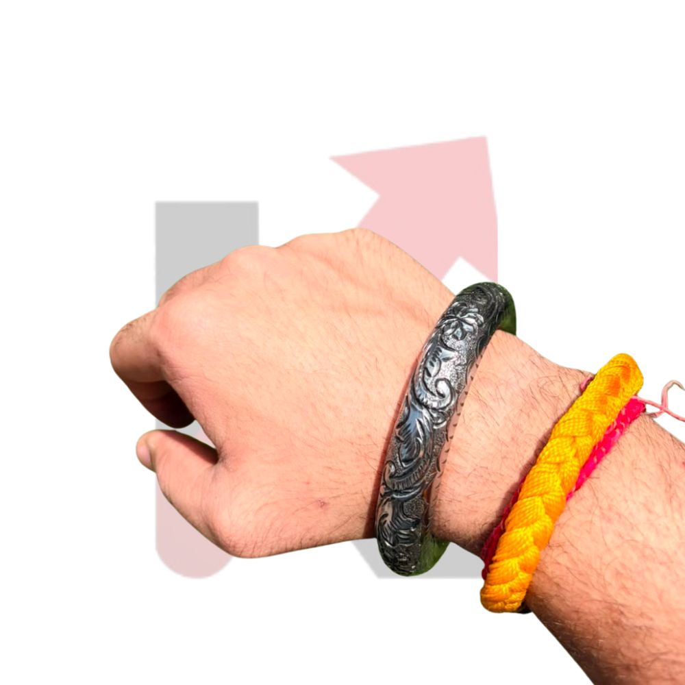 A close-up of a person's wrist wearing a dark engraved metal bracelet, with two colorful braided cord bracelets (yellow and pink) on the same wrist, set against a plain white background with faint pink and gray arrow graphics.