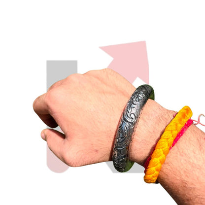 A close-up of a person's wrist wearing a dark engraved metal bracelet, with two colorful braided cord bracelets (yellow and pink) on the same wrist, set against a plain white background with faint pink and gray arrow graphics.